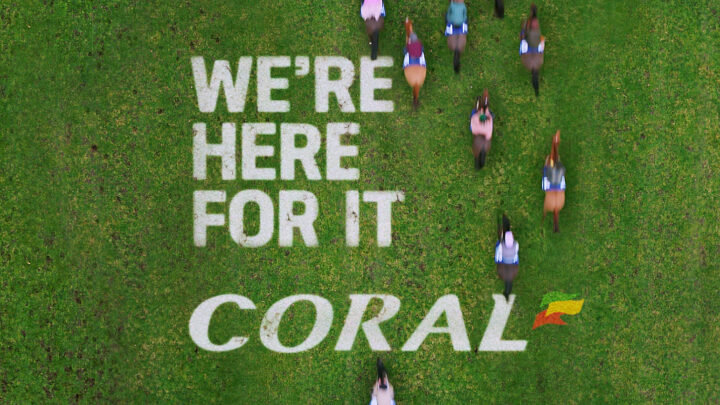 Birds eye shot of the race track. The words 'We're Here For It Coral' in white on the floor, with horse running over it.