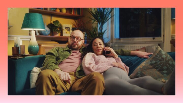 couple sits on a sofa watching TV, the wife taking a phone call while the husband looks on annoyed